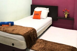 A hotel room with a single bed, a brown blanket, an orange pillow with "KOOL KOST" logo, a white pillow, and a small shelf with a potted plant.
