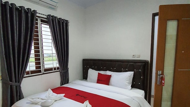 A hotel room with a bed, a window with curtains, an air conditioning unit, and a door. - RedDoorz near CitraLand Gama City Medan (Medan)