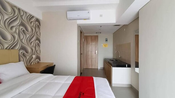 Habitación de hotel con cama, mesita de noche, colcha roja, pared con diseño ondulado, zona de cocina con lavaplatos y armarios, y unidad de aire acondicionado.