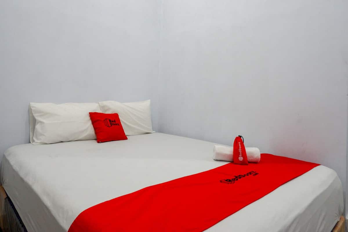 A neatly made bed with white bedding and a red blanket, a red pillow, and a red towel with a logo.