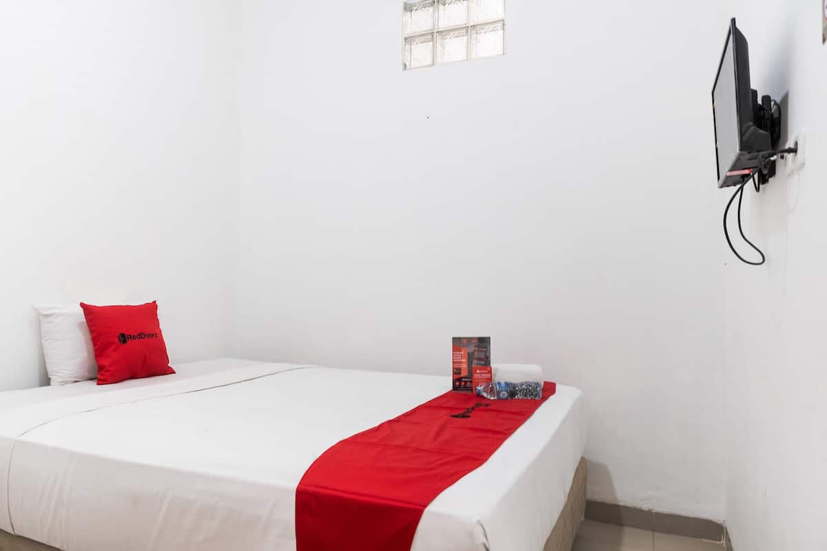 Habitación individual con ropa blanca de cama, una colcha roja, un cojín rojo, un televisor montado en la pared y una ventana pequeña.