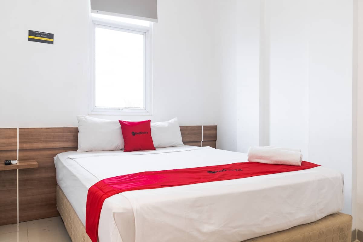 Habitación de hotel con cama, ventana, cabecero de madera, almohada roja, toalla blanca y un estante pequeño.