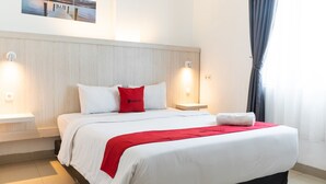 Una cama bien hecha con ropa de cama blanca y roja, dos mesitas de noche con lámparas y vistas al exterior a través de una ventana con cortinas.