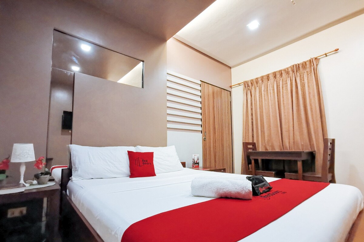 A hotel room with a large bed, a desk with a lamp, a chair, and a window with curtains.