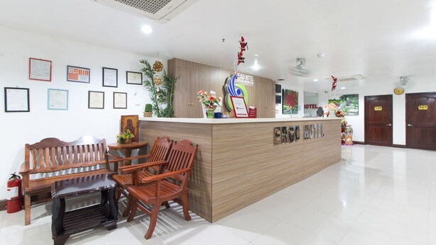 A hotel reception area with a wooden reception desk, framed certificates on the wall, and a seating area with wooden chairs and a bench. - RedDoorz Plus near Vigan City Commercial Center (Vigan)