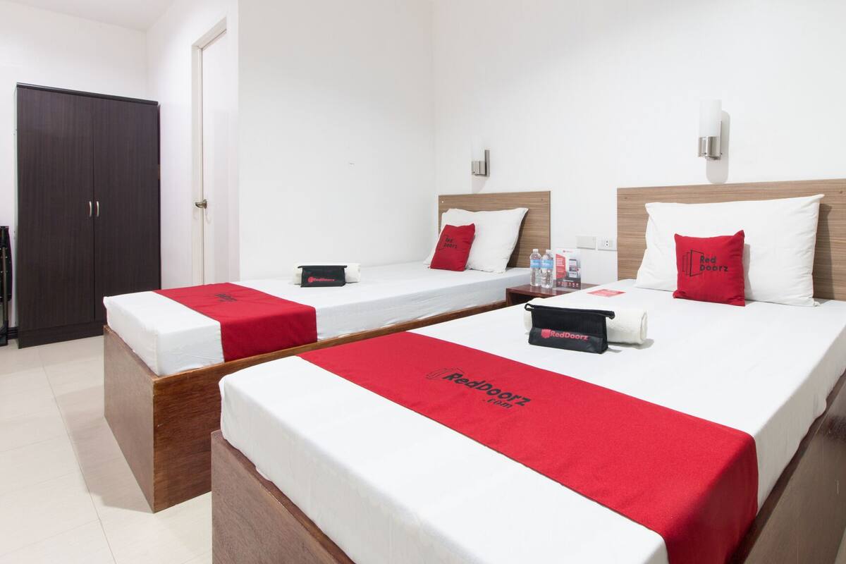 Habitación de hotel con dos camas, cada una con colcha y almohadas rojas, cabecero de madera y una mesita de noche con una botella y una caja de pañuelos.