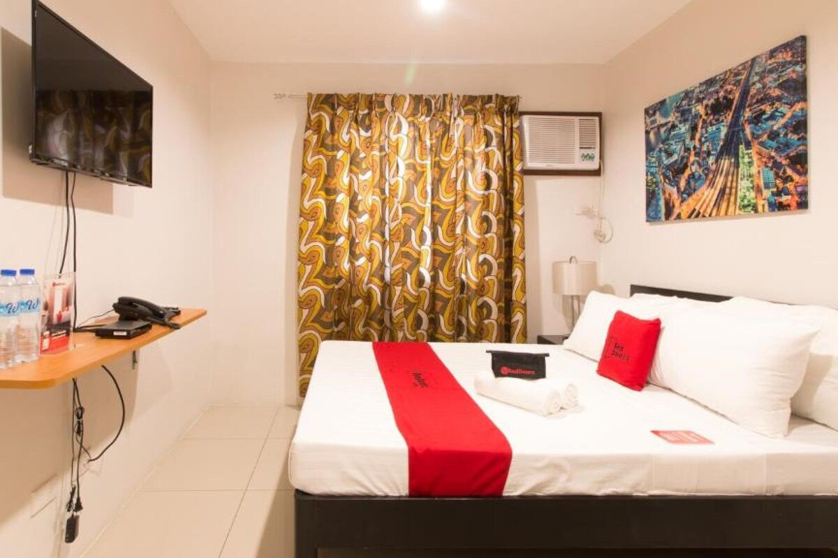 A hotel room with a bed, a desk with a phone and water bottles, a TV mounted on the wall, and a colorful abstract painting above the bed.