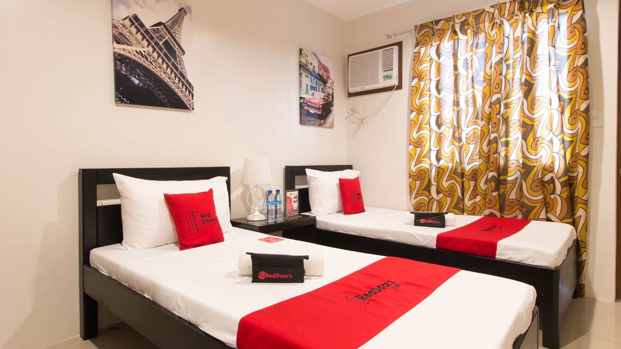A hotel room with two beds, each with white bedding and red accents, including pillows and a blanket. There are wall art pieces depicting the Eiffel Tower and a cityscape. A window with patterned curtains is visible.