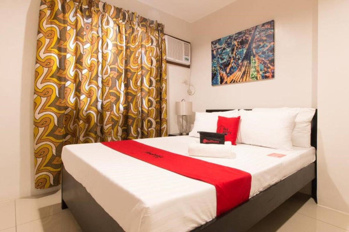 Habitació d'hotel amb un llit, una manta vermella, un quadre a la paret i cortines estampades.