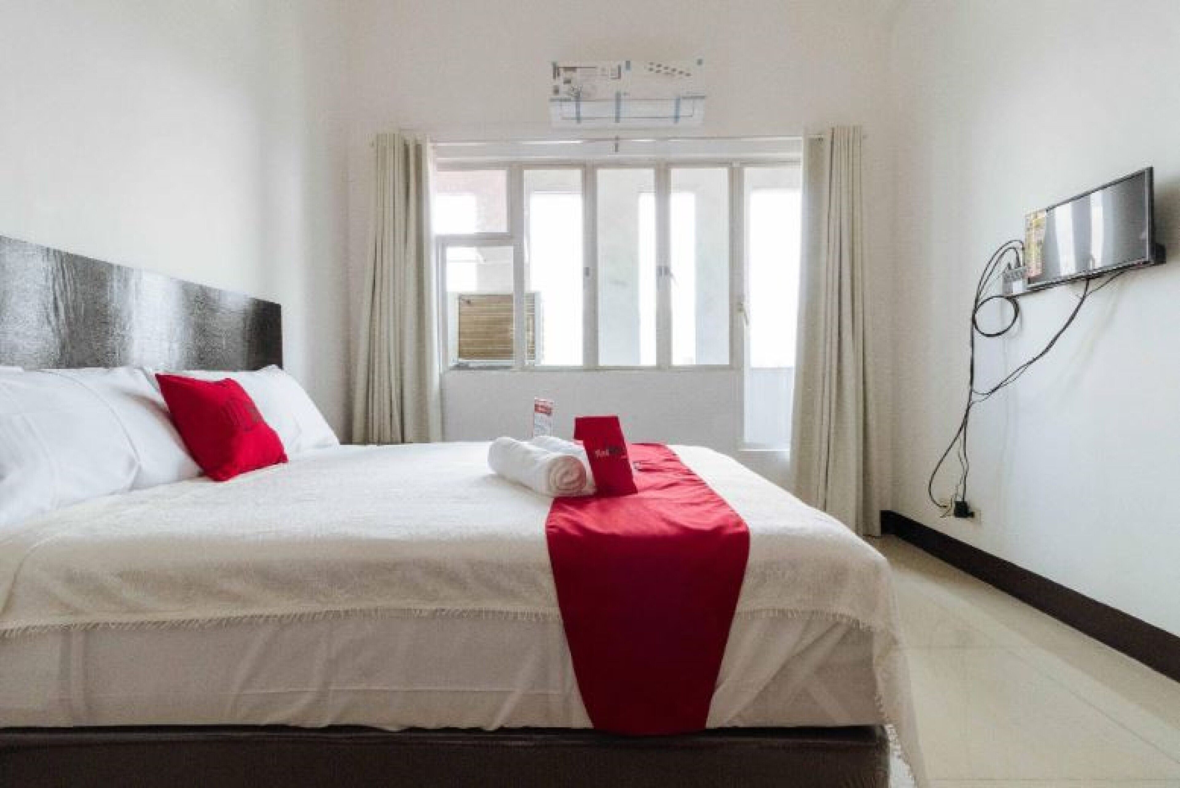 Habitación de hotel con cama, televisor montado en la pared y ventana con cortinas.
