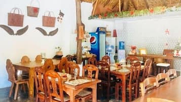 Uma área de refeições com mesas e cadeiras de madeira, teto de palha e uma parede decorada com cestos e arte.