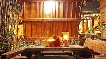 Uma mesa de madeira com uma vela, uma garrafa e um copo em ambiente rústico ao ar livre.