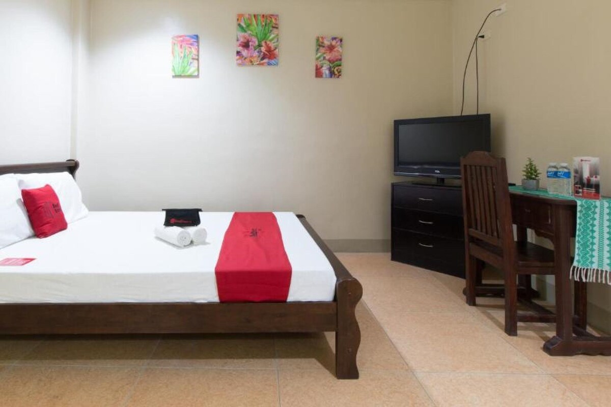 Una cama con una manta roja, un televisor y un escritorio con una silla.
