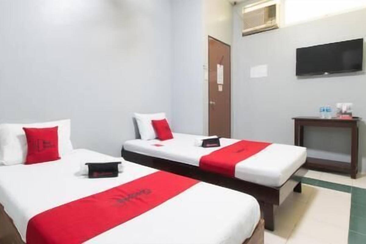 A hotel room with two beds, each with white bedding and red accents, a small bedside table, a television mounted on the wall, and a door in the background.