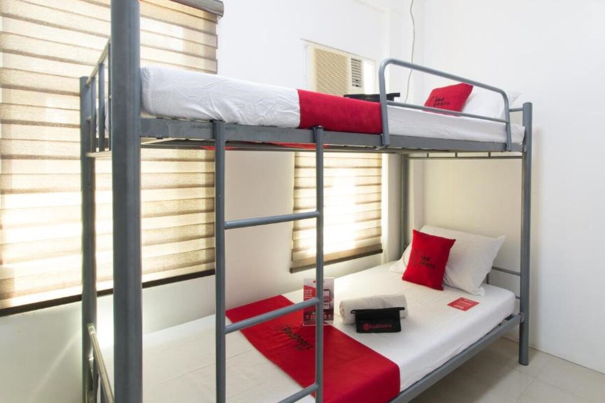 Una cama individual con ropa de cama roja y blanca, una mesita de noche pequeña y una ventana con persianas.