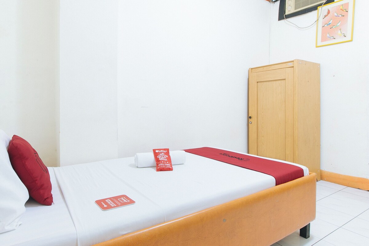 Ein Einzelbett mit Holzrahmen, weiße Bettwäsche, ein rotes Kissen und ein rotes Handtuch mit einem Logo. An der Wand steht ein Holzkasten und darüber hängt ein gerahmtes Bild.