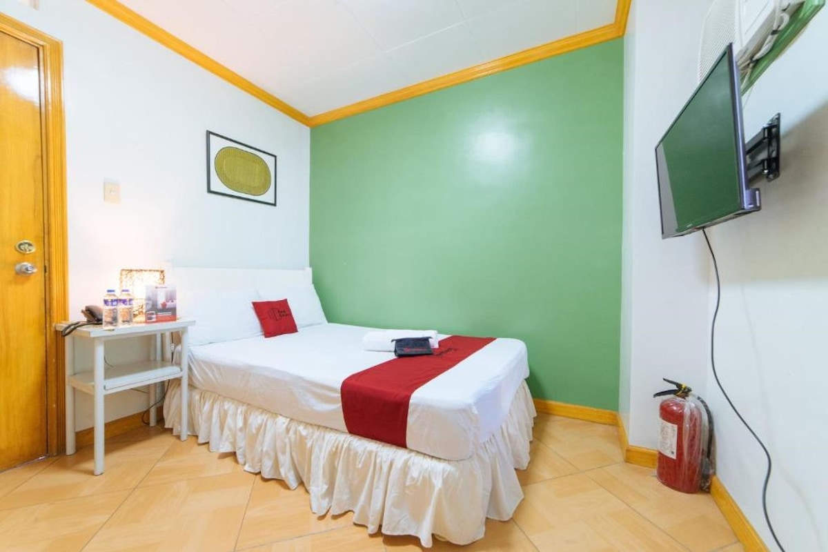 A single bed with a red and white bedspread, a bedside table with a lamp, a television mounted on the wall, and a fire extinguisher.