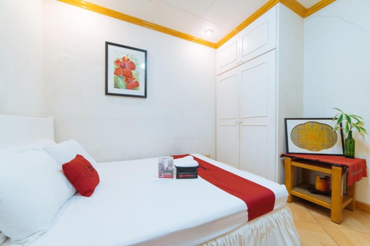 A neatly made bed with a red pillow, a framed picture on the wall, and a small table with a plant.