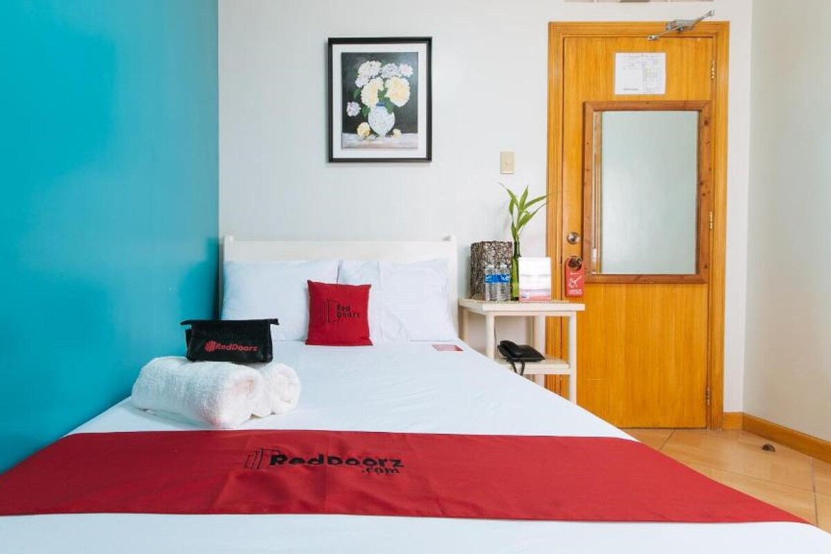 A hotel room with a bed, a red blanket, a pillow, a framed artwork, a bedside table with a plant, and a wooden door.