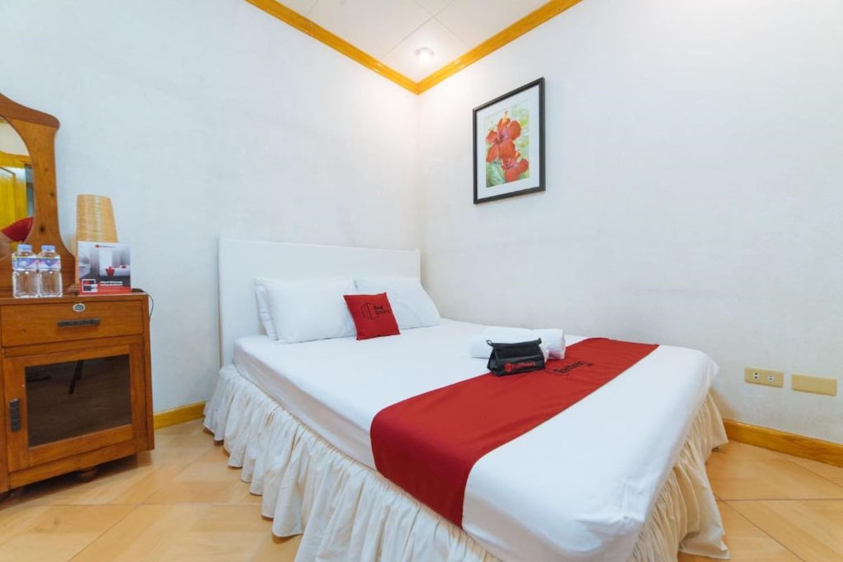 Habitación de hotel con cama, mesita de noche de madera con lámpara, almohada roja y un cuadro en la pared.