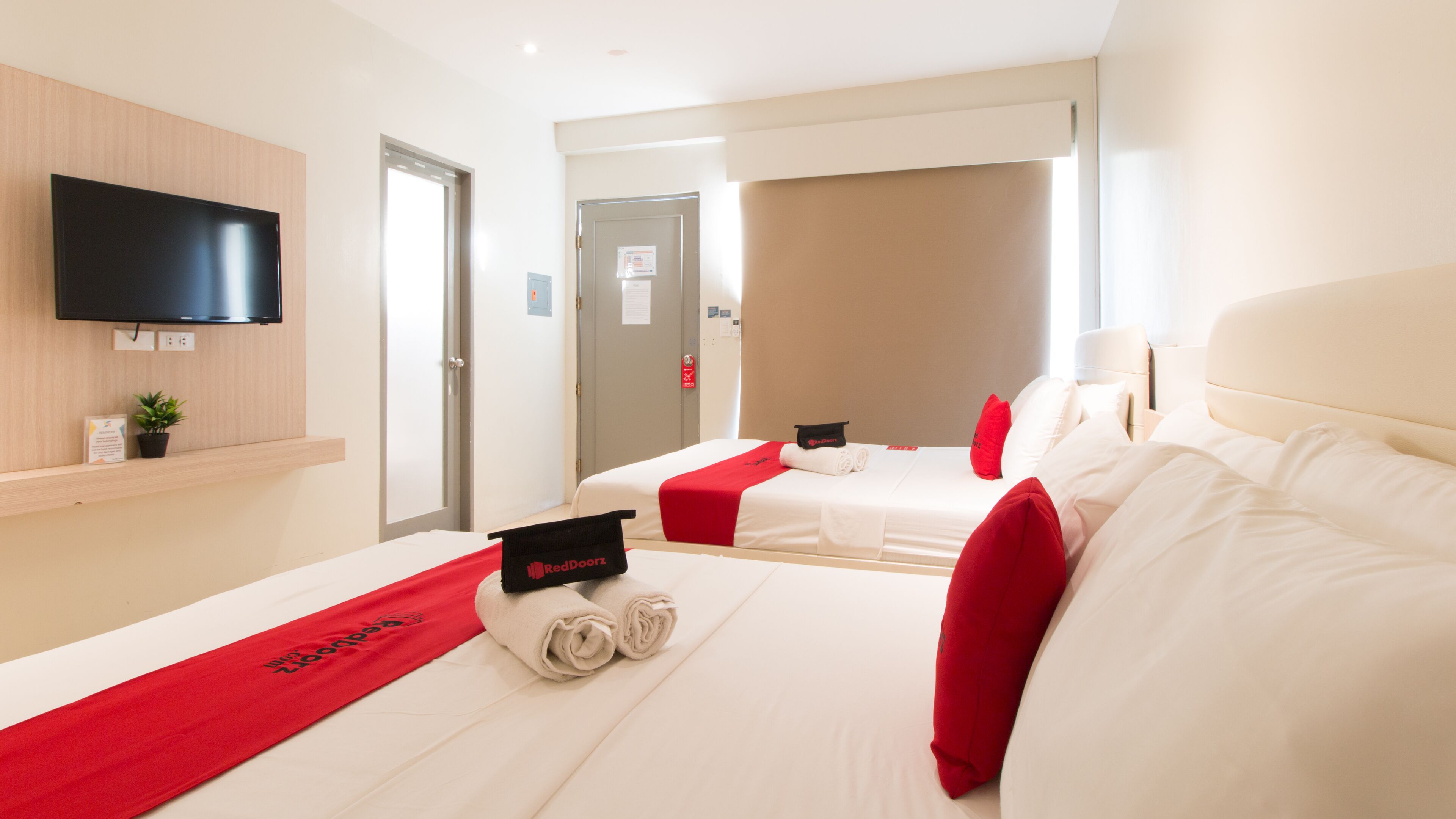 A hotel room with a flat-screen TV mounted on the wall, a bed with white bedding and red accents, and a red towel with the RedDoorz logo.