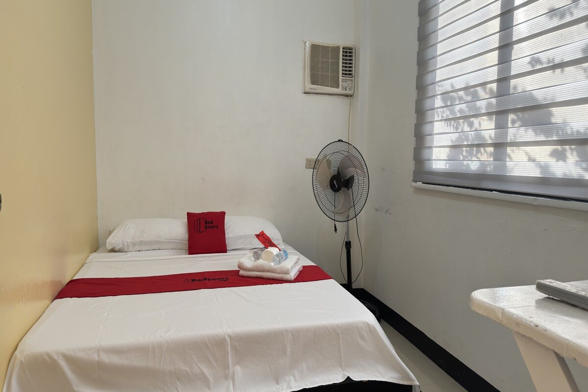 A small, clean room with a bed, a bedside table, a fan, and a window with blinds.