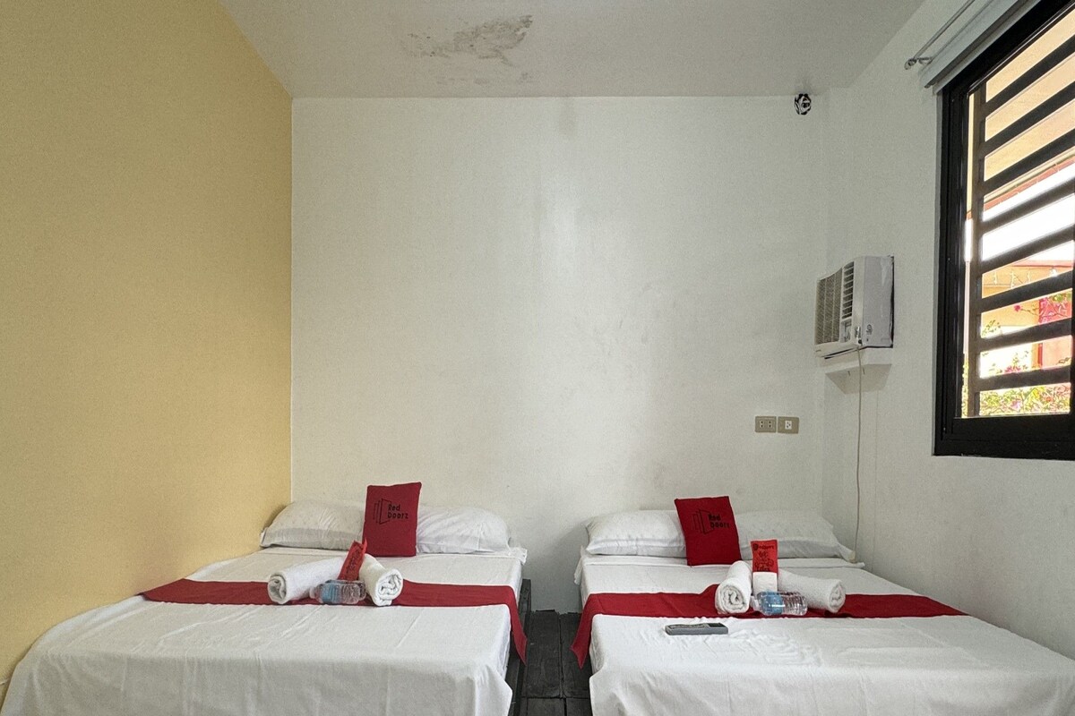 A hotel room with two single beds, each with a red pillow and a white bedspread. There is a window with blinds on the right side, and an air conditioning unit mounted on the wall.