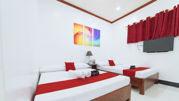 Une chambre d’hôtel avec deux lits simples, chacun agrémenté de draps blancs et de touches de rouge, d’un plaid rouge et d’un oreiller rouge. On y trouve une petite table avec une lampe et une bouteille de lotion. La chambre a des murs blancs, un plafond en bois et un rideau rouge à la fenêtre.