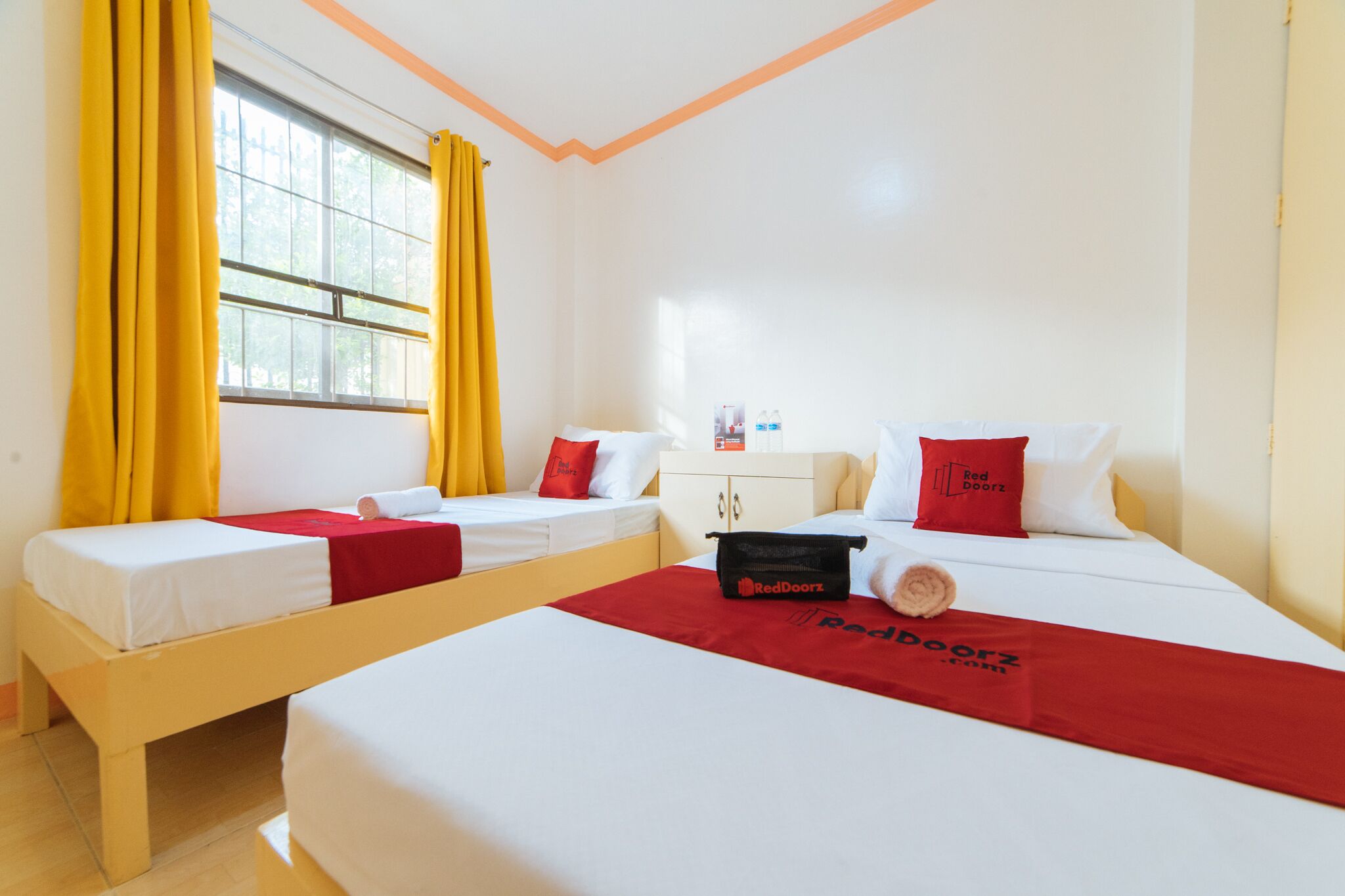 A hotel room with two beds, yellow curtains, a small table with a water bottle, and a RedDoorz bag on one bed.