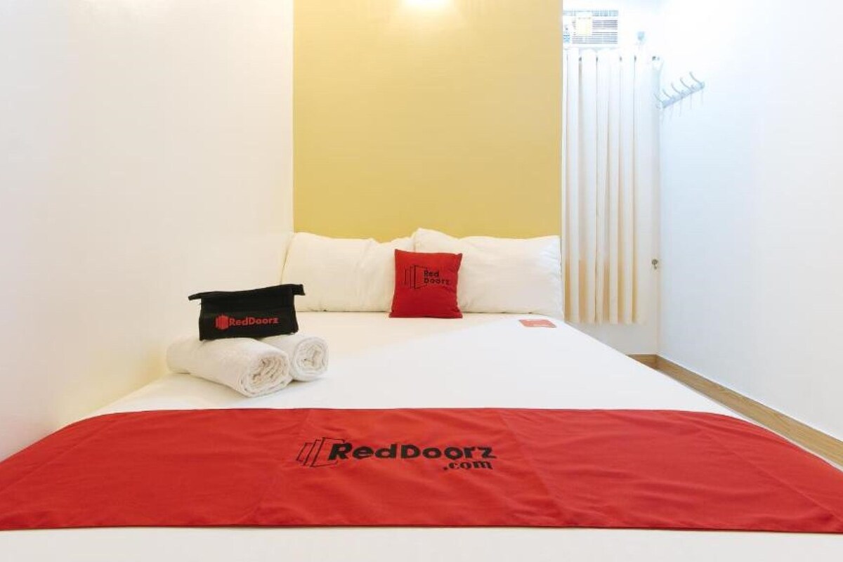 A hotel room with a neatly made bed, a red blanket, and a RedDoorz logo on the bedspread.