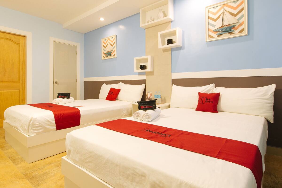 A hotel room with two beds, each with white bedding and red accents, a wooden door, and a light blue wall with decorative elements.