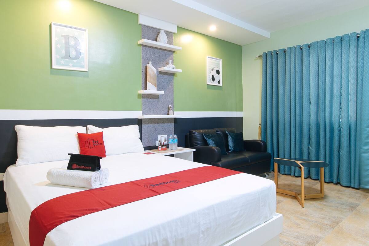A hotel room with a bed, a nightstand, a chair, a sofa, a wall with a shelf, and a window with curtains.