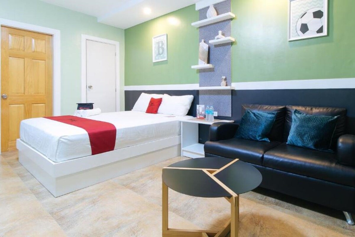 A bedroom with a bed, a black leather sofa, a small table, and a chair. There are shelves with decorative items and a door.