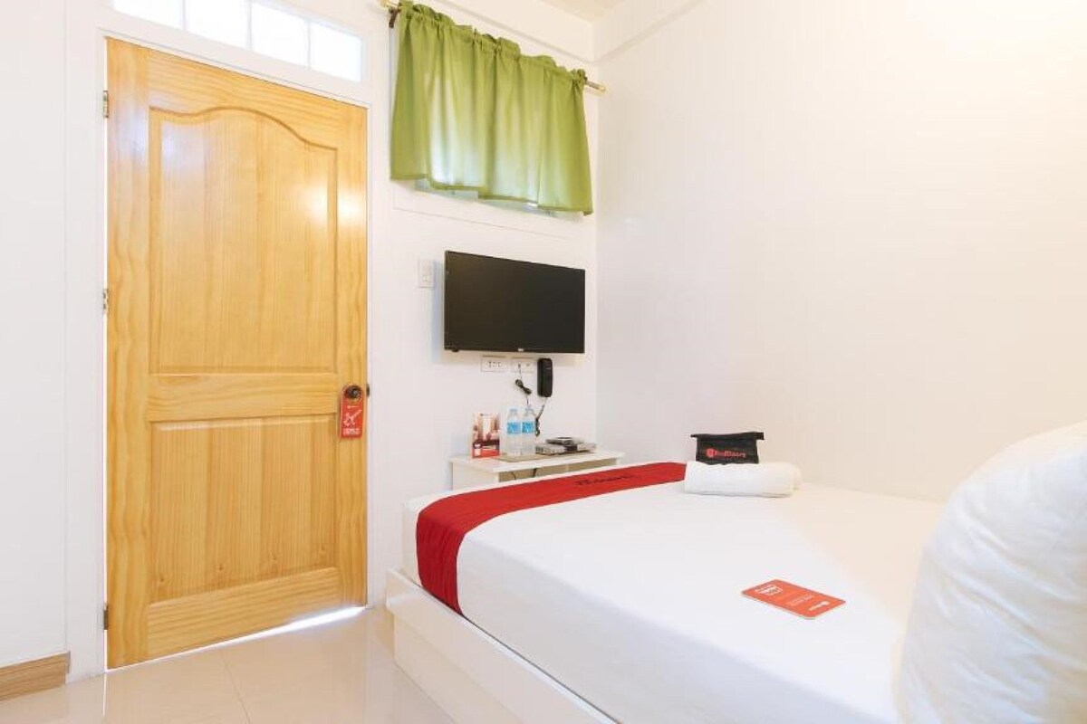 A hotel room with a wooden door, a flat-screen TV mounted on the wall, a small bedside table with a bottle and a red object, and a bed with a white mattress and red bedding.