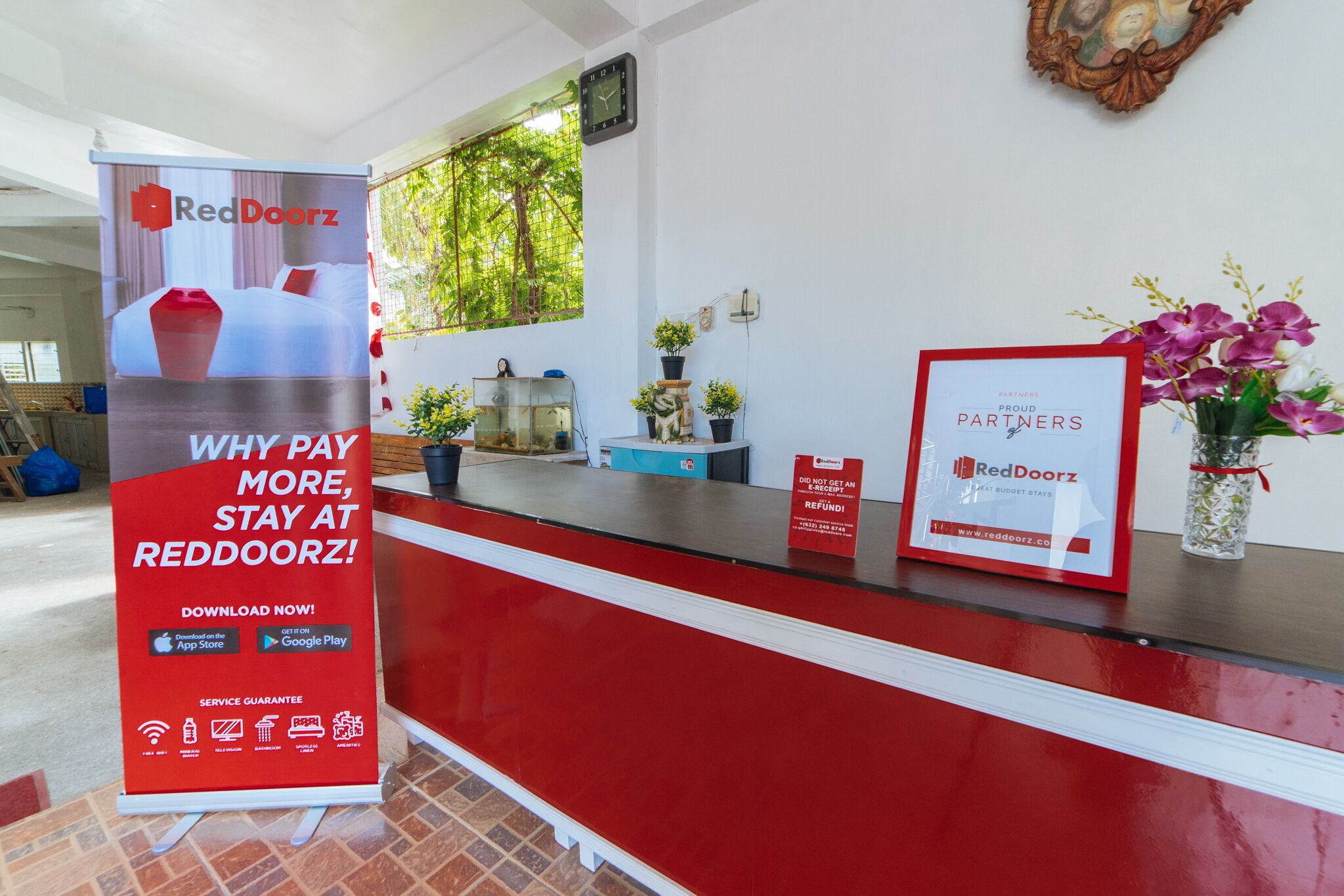 A hotel reception desk with a RedDoorz sign, a framed certificate, and a vase with flowers.