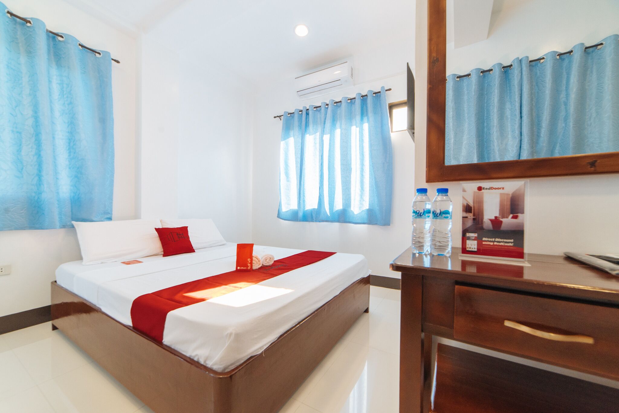 A hotel room with a bed, a desk, and a window with curtains. There are water bottles on the desk.
