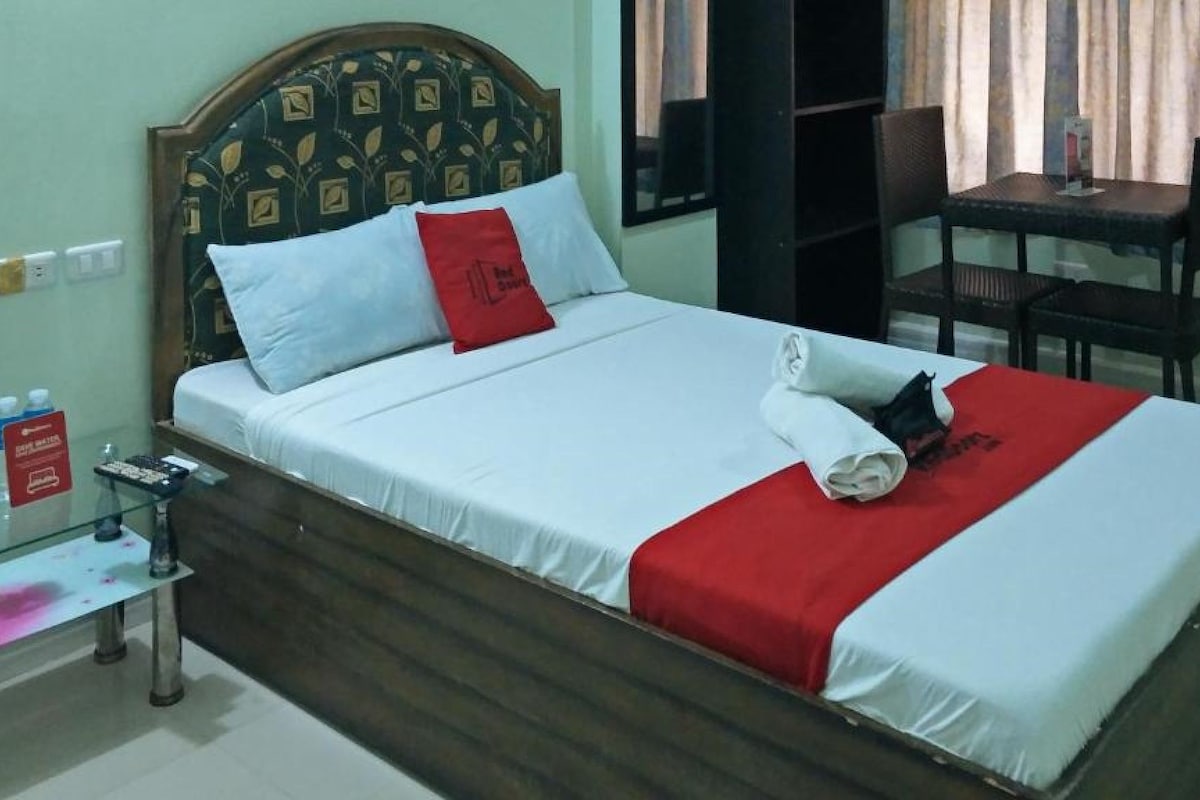 A neatly made bed with white and red bedding, a wooden headboard with a leaf pattern, a small table with a chair, and a bottle of hand sanitizer on a glass table.
