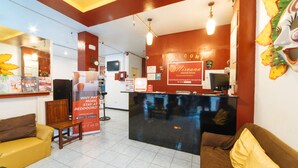 Un espace d’accueil comprenant un comptoir noir, un canapé marron, un canapé jaune et un mur rouge orné d’une pancarte de bienvenue.