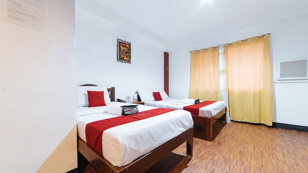 Une chambre d’hôtel avec deux lits, un plancher en bois, une couvre-lit rouge et blanche, un oreiller rouge, une petite table recouverte d’une nappe rouge et une fenêtre avec des rideaux jaunes.