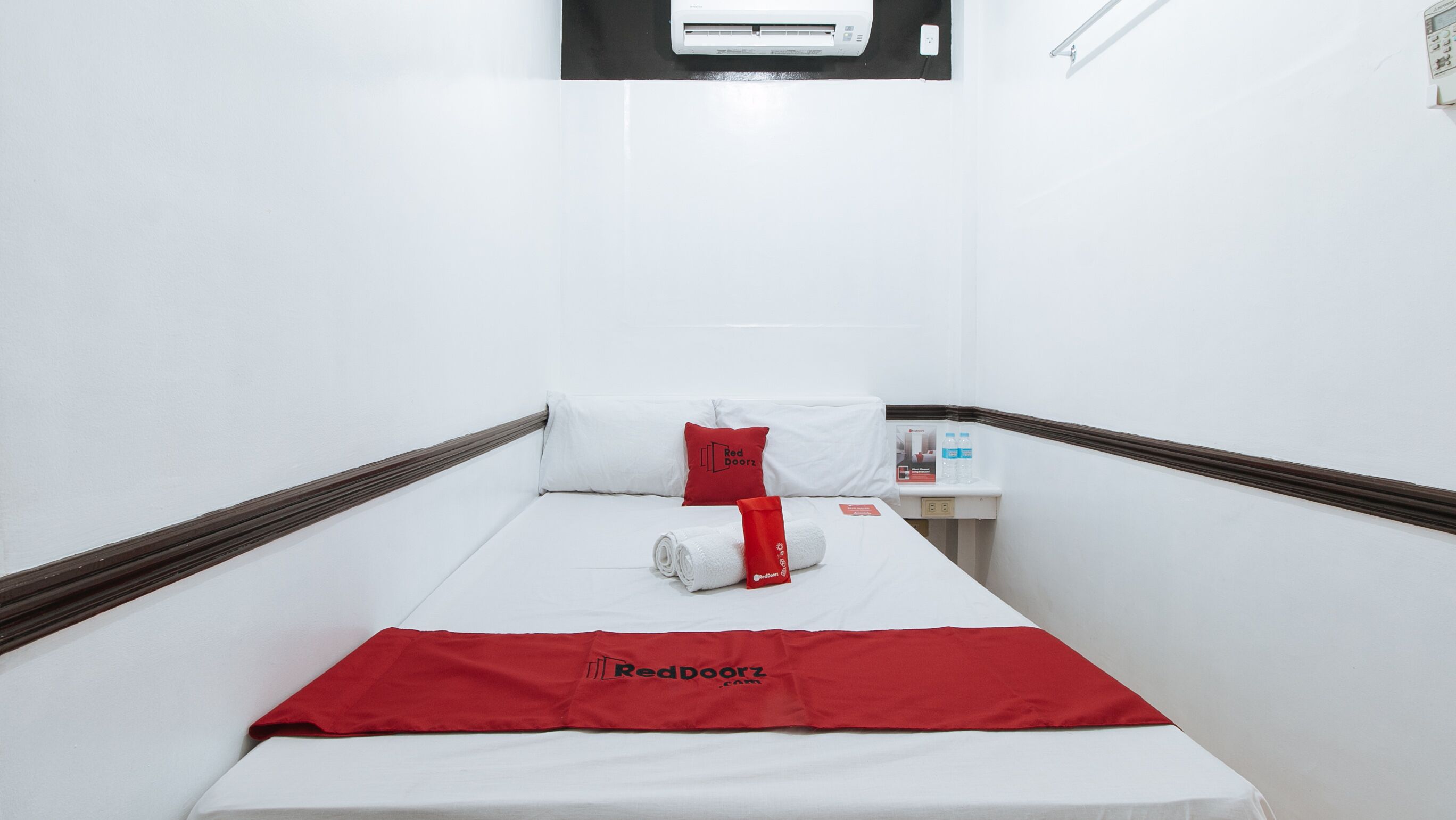 A hotel room with a bed, a red blanket, a pillow with the RedDoorz logo, a water bottle, and a small table with a bottle of water.