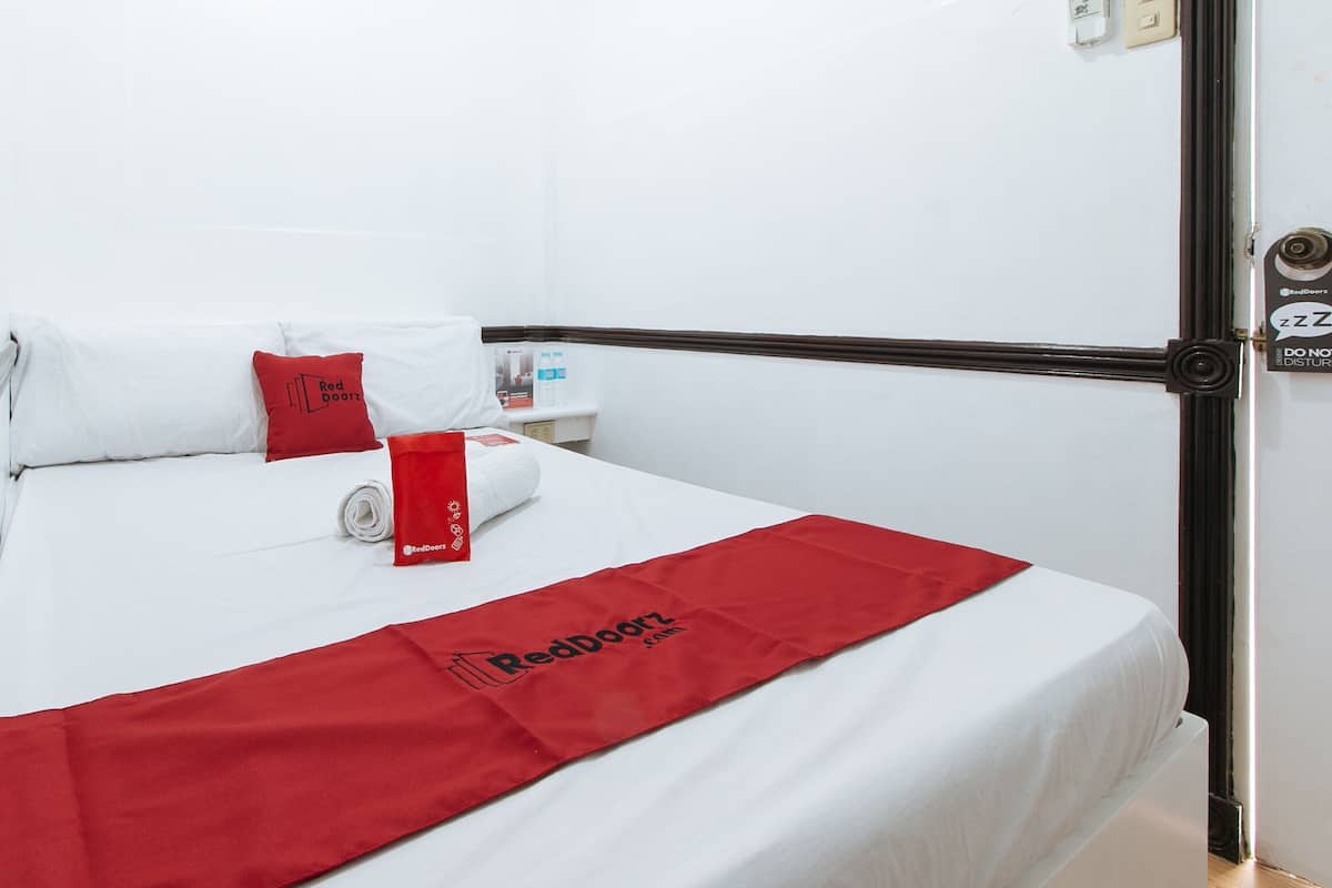 A hotel room with a bed, white bedding, a red blanket, a red pillow, a red bag, and a water bottle on the nightstand.