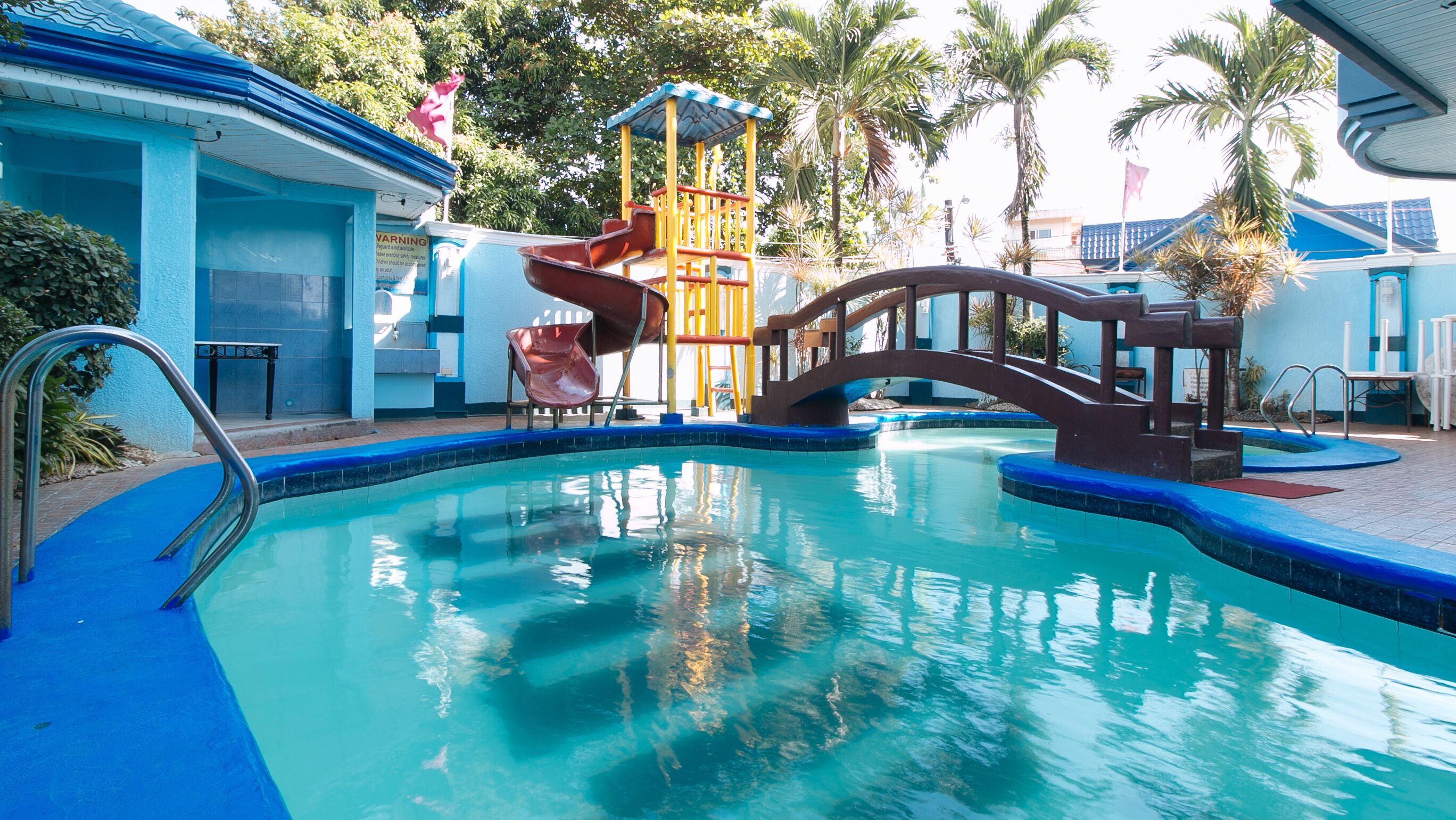 A swimming pool with a bridge and slides, surrounded by palm trees and a building with blue walls.