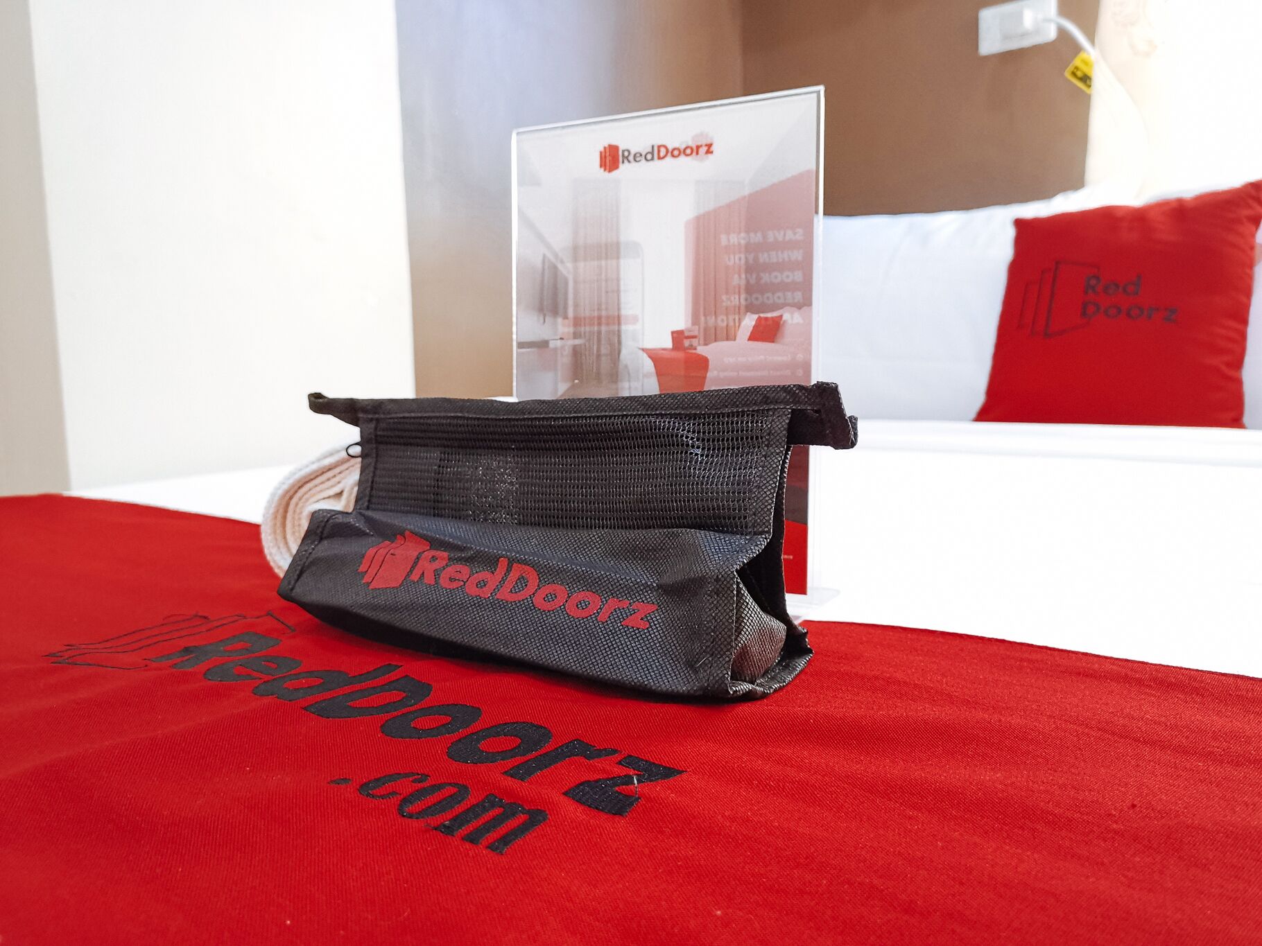 A RedDoorz branded bag on a red bedspread with the website 'RedDoorz.com' visible.