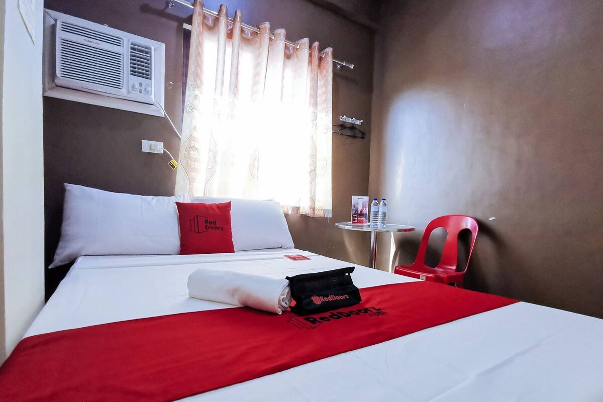 Habitación de hotel con una cama, una mesita con botellas de agua, una silla roja y un equipo de aire acondicionado.