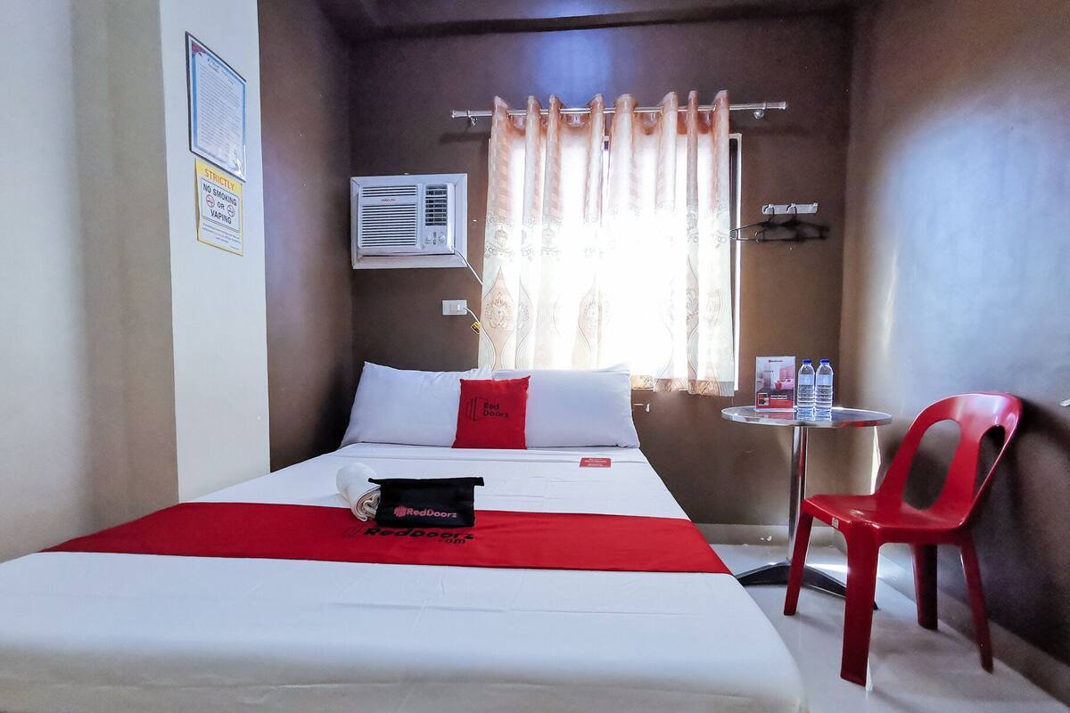 Habitación de hotel con cama, una mesita con dos botellas, una silla roja y una ventana con cortinas.