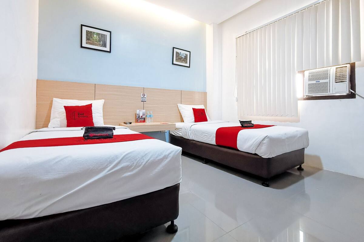 A hotel room with two beds, each with a red and white bedspread, a wooden headboard, and a small table with a red pillow and a Red Doorz bag.