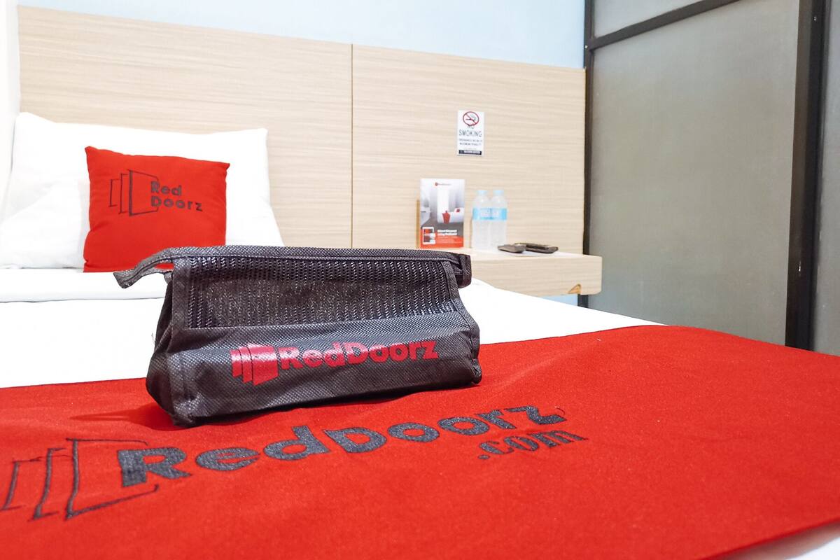 A hotel room with a bed, a red pillow, a Red Doorz bag, a water bottle, and a no-smoking sign on the wall.