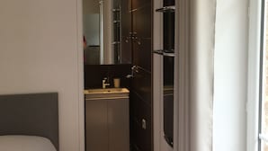 Ein schmales Zimmer mit Bett, kleinem Badezimmer und einem Fenster mit Vorhängen.