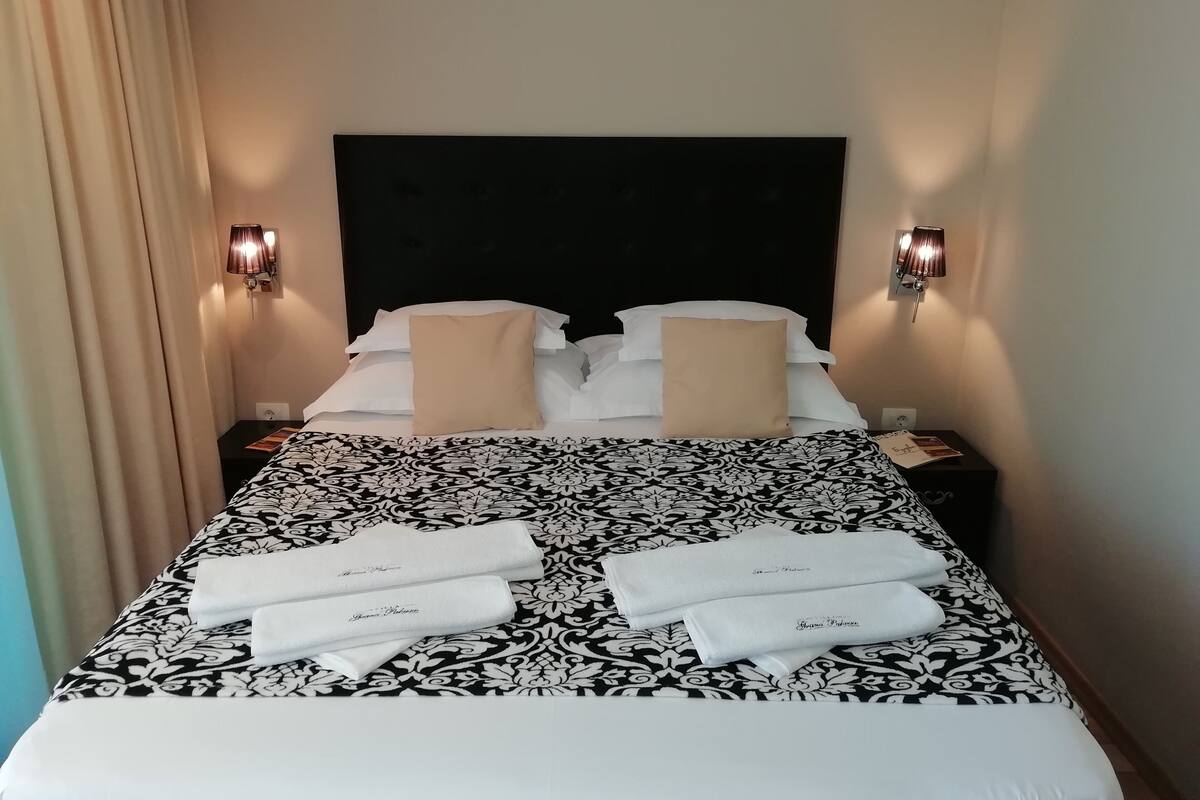 Una cama bien hecha con colcha de rayas blanco y negro, dos almohadas beige y dos toallas blancas con texto. Hay dos lámparas de pared a cada lado de la cama.