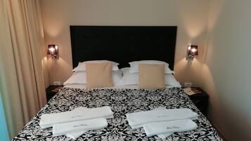 A neatly made bed with a black and white patterned bedspread, two beige pillows, and two white towels with text on them. There are two wall-mounted lamps on either side of the bed.